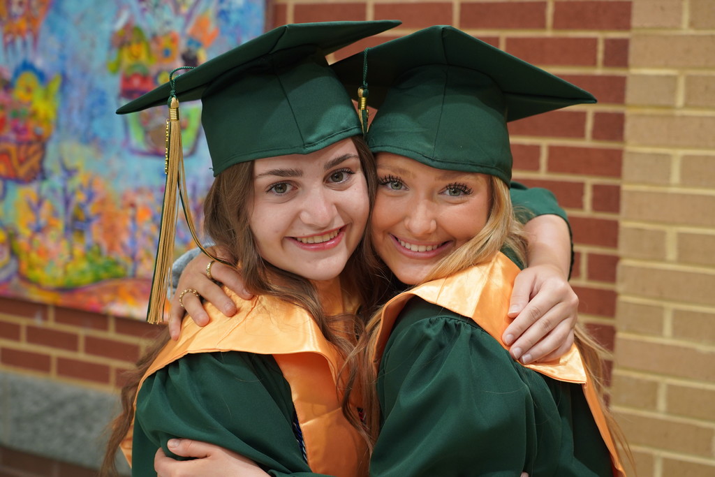 Two women, in green gowns and caps, stand close together, smiling at a brick wall.