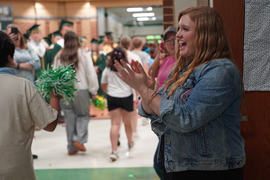 A woman in a denim jacket celebrates, clapping, as a person with a pom-pom stands nearby.