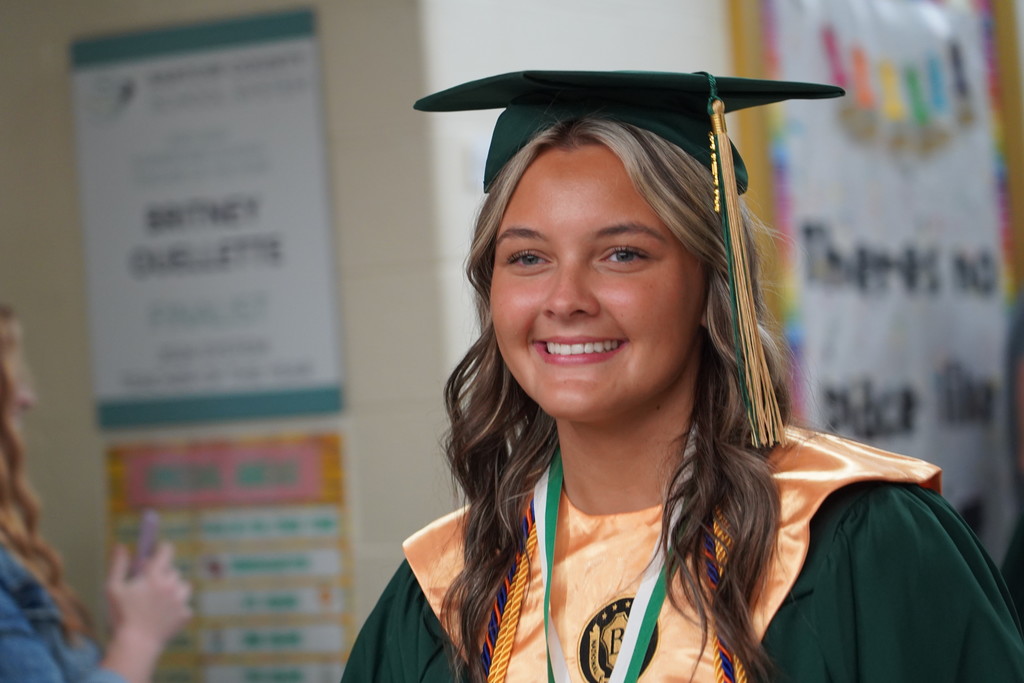 Woman in a graduation gown with tassel smiles for the camera, with educational posters behind.
