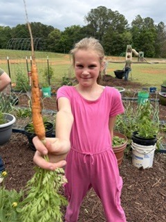 Girl holding a carrot in a garden with greenery and trees in the background.