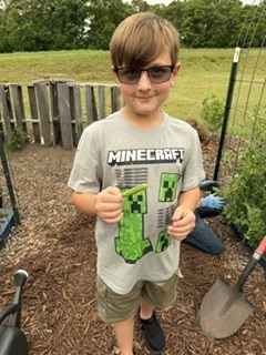 A boy in a Minecraft t-shirt holds a plant in a garden with wooden fence and green grass.