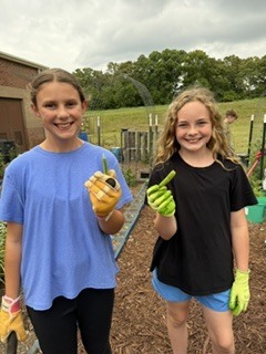 Two girls in gloves hold green plants in a garden with a building, greenery, and a cloudy sky.