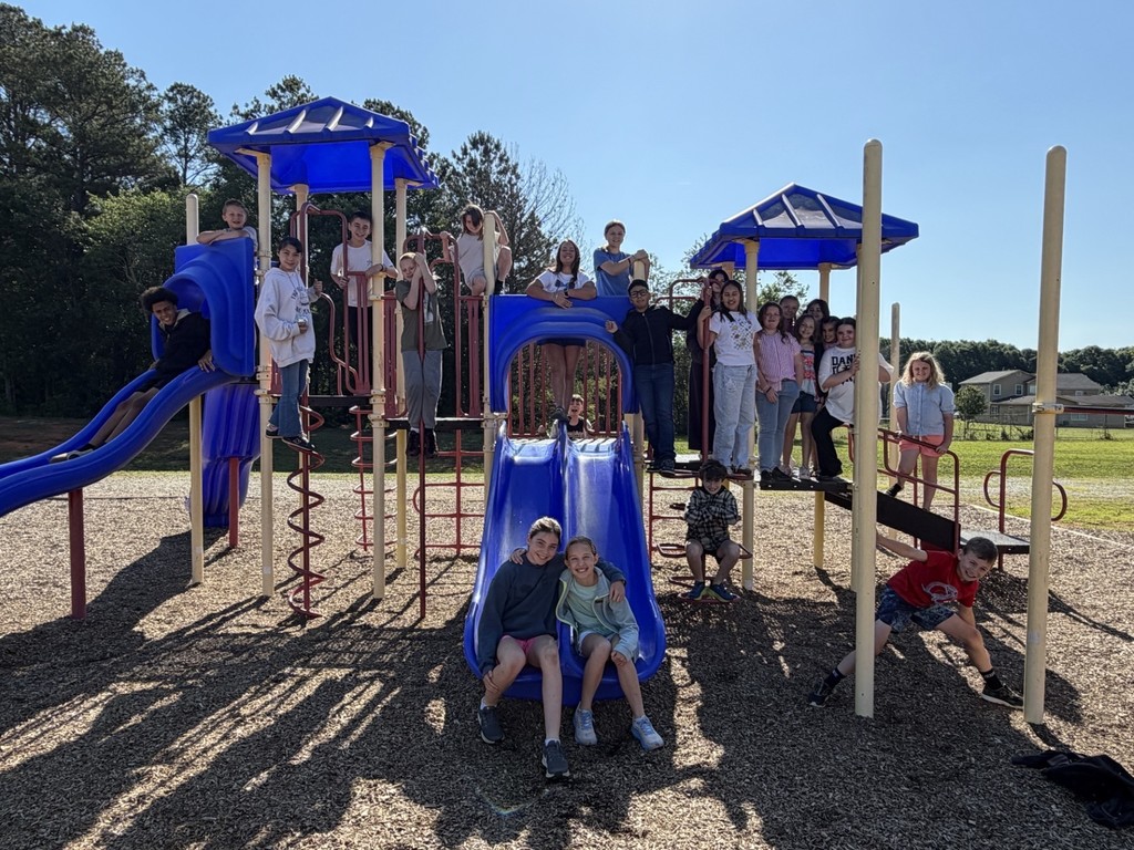 Group of children on playground with slides and play equipment under a blue sky with trees in background.