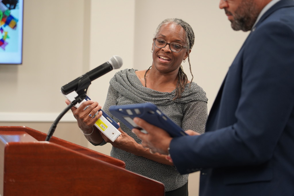 A woman with glasses holds a book while standing at a podium. A man in a suit examines a blue folder.