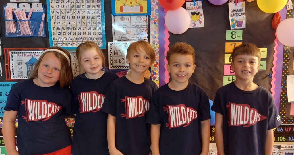 Five children in matching blue shirts with "Wildcats" text stand together in front of a classroom wall with balloons.