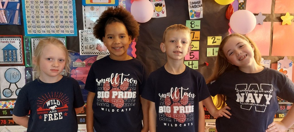Four children in navy t-shirts pose for a photo against a vibrant, colorful classroom backdrop.  The shirts says "Wild and Free", "Small Town Big Pride Wildcats", and "Wildcat Wednesday Navy".
