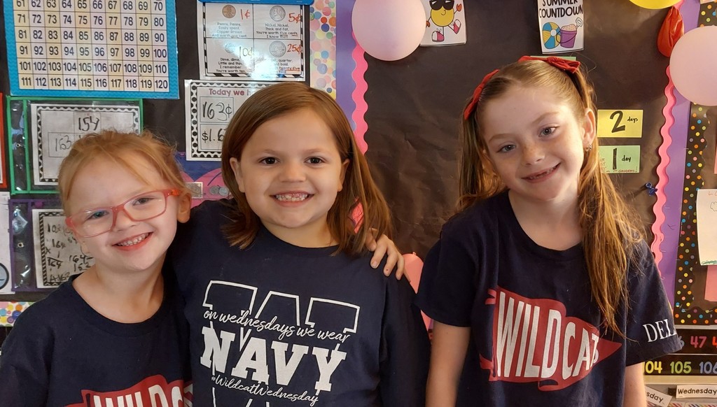 Three girls in matching shirts stand in front of a classroom bulletin board with balloons.