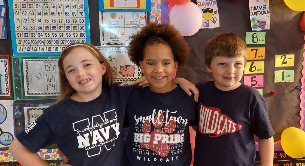 Three children wearing navy blue shirts stand together, smiling, in front of a bulletin board.