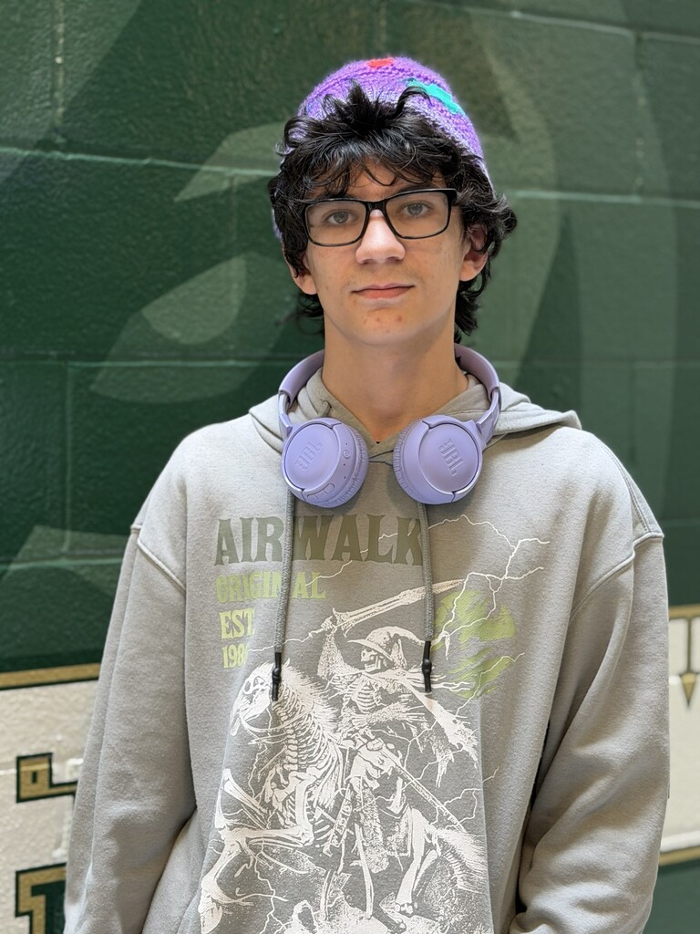 A young person with glasses and a purple hat stands against a green wall. They wear a gray hoodie with an Airwalk design and purple headphones around their neck.