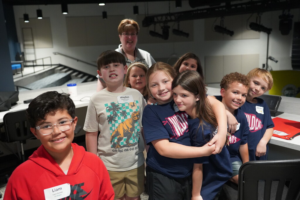 Nine children in matching shirts, smiling and posing for a group photo. Behind them, a woman stands near a staircase.