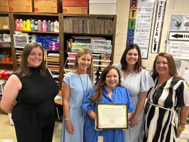 Five women stand in a classroom. A central woman holds a framed certificate. Behind them, shelves hold various items. A bulletin board with text is visible.