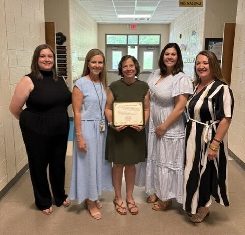 Five women stand in a hallway, one holding a framed certificate. They wear dresses and sandals, smiling at the camera. The hallway has white walls and a tiled floor.
