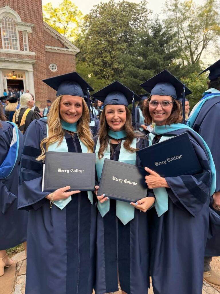 Three women in academic regalia hold diplomas in front of a building with a crowd in background.