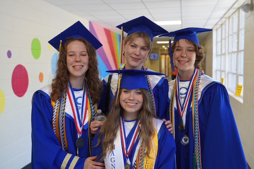 Four students in blue caps and gowns, possibly for graduation, standing close together in a hallway.
