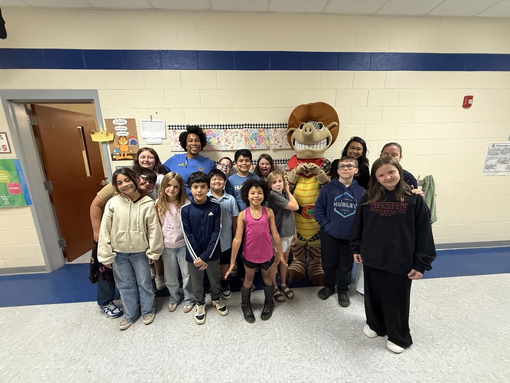 Group of students and staff posing for a photo with a mascot in a classroom with white walls and blue trim.