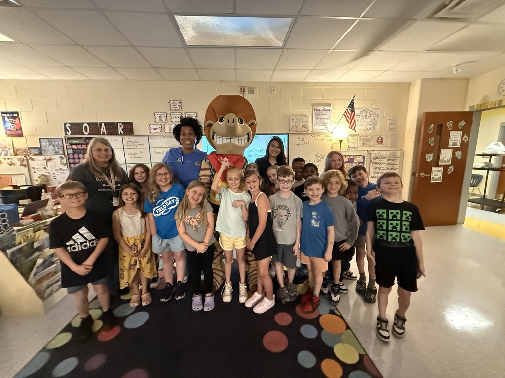 A classroom with a carpet and a mascot. A group of kids, a woman, and a man stand together.