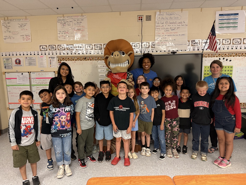 A group of children in a classroom with a mascot in front of a bulletin board and an American flag.