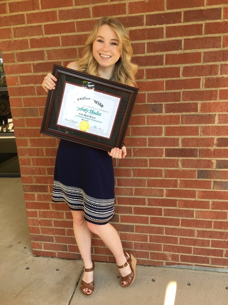 A woman in a navy dress, holding a framed certificate, smiles in front of a red brick wall.