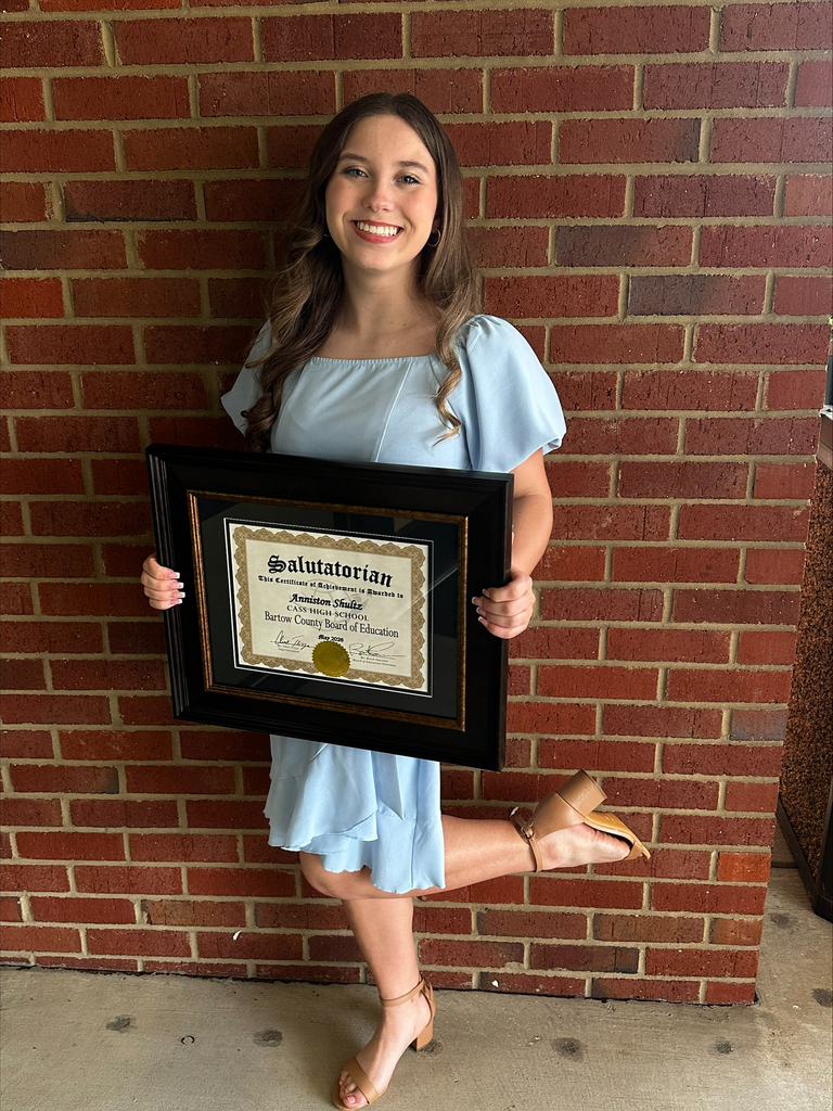 A woman in a light blue dress holds a framed certificate, smiling in front of a red brick wall.