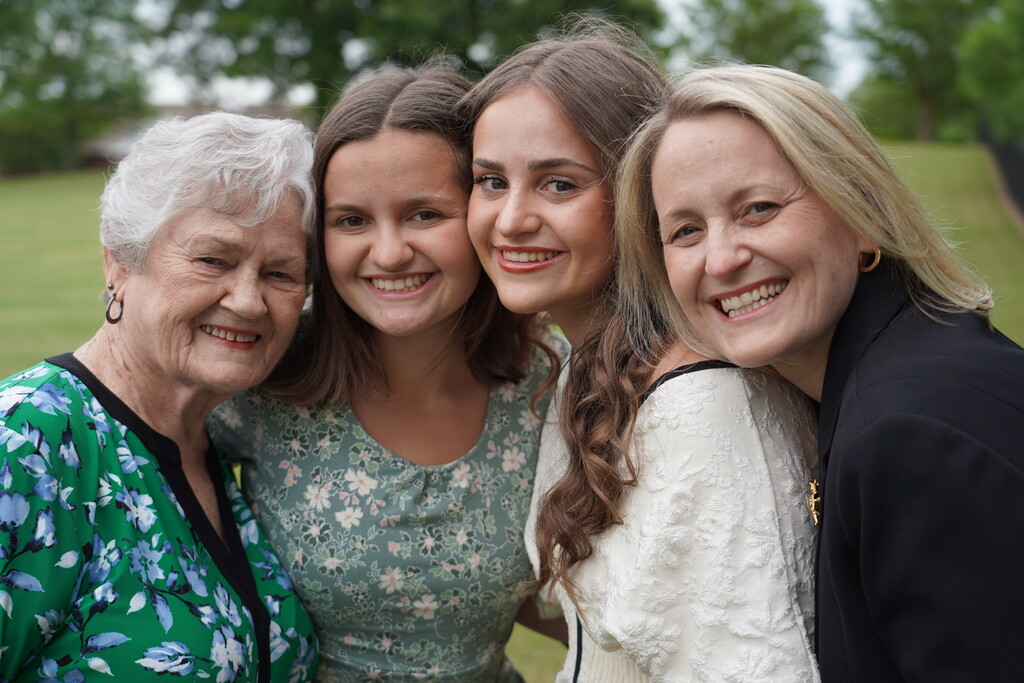 Five women smile in a park with trees and a building in the background.