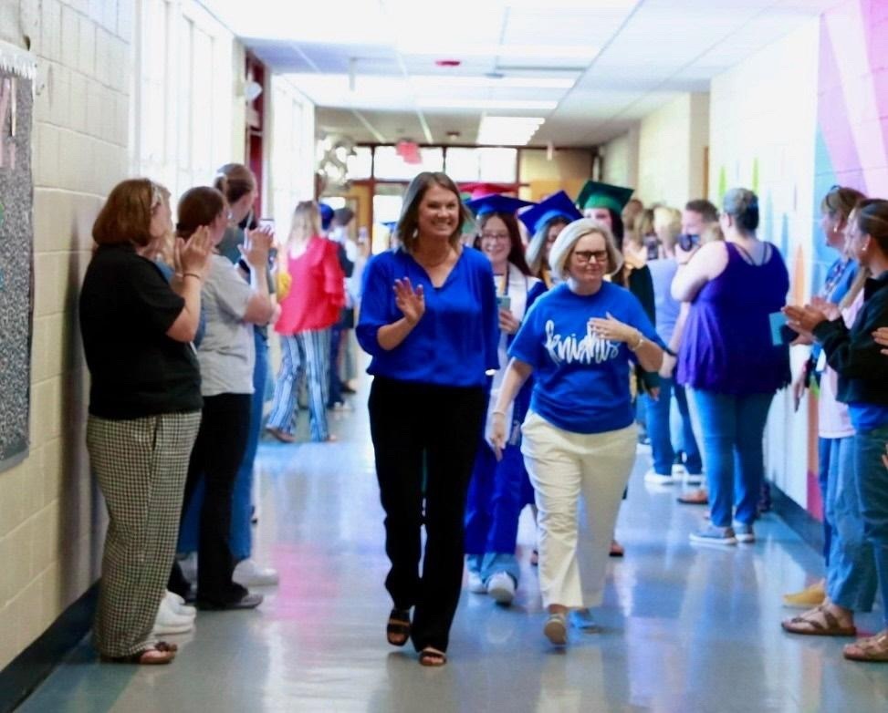 A group of people wearing blue shirts and caps walk down a hallway, lined with walls and pillars, as others stand by to cheer them on.