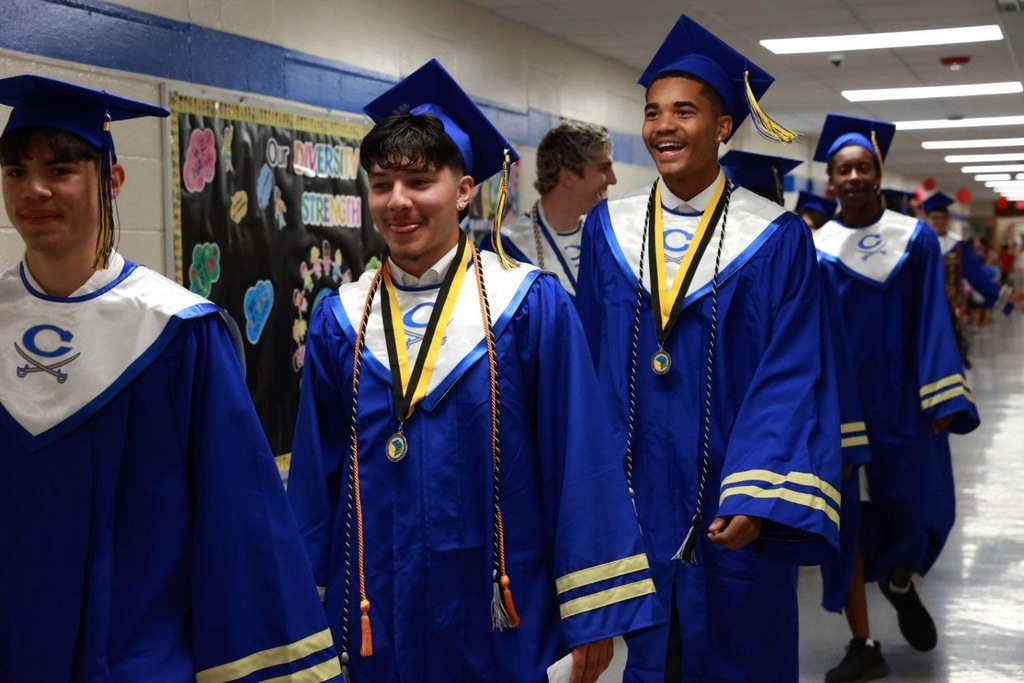 Group of students in blue graduation gowns and caps walk down a school hallway with artwork on the wall.