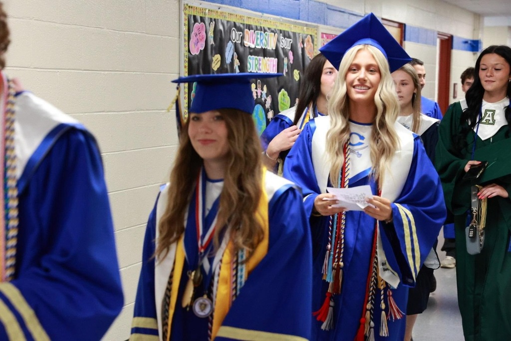 Five students in blue and gold gowns and caps walk down a hallway. One holds a piece of paper.