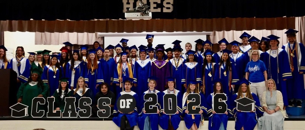 Graduates in blue gowns and caps stand on stage with text "CLASS OF 2026". A white backdrop is behind them.