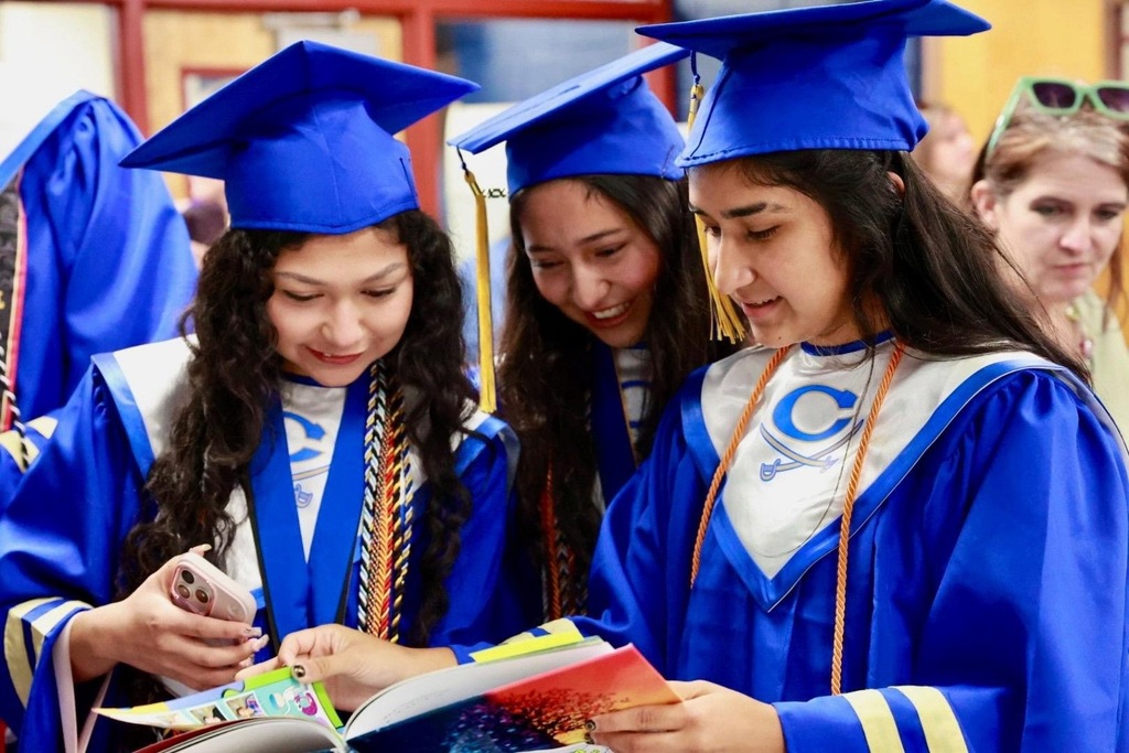 Four students in blue gowns and hats are looking at a book, smiling and laughing.
