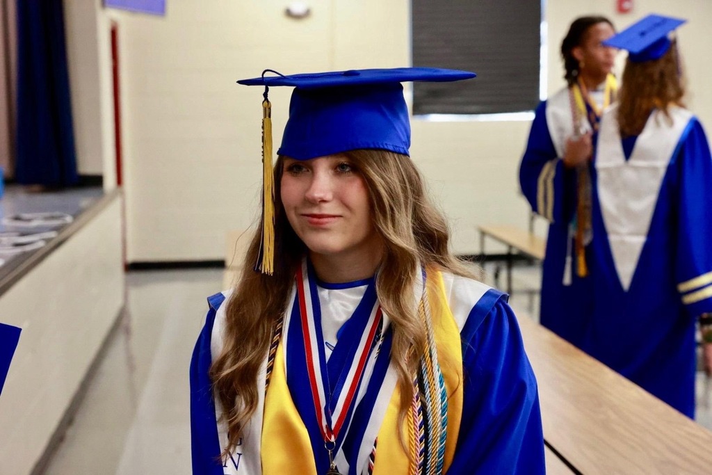 A young woman in blue and gold graduation attire stands in a hallway with other students.