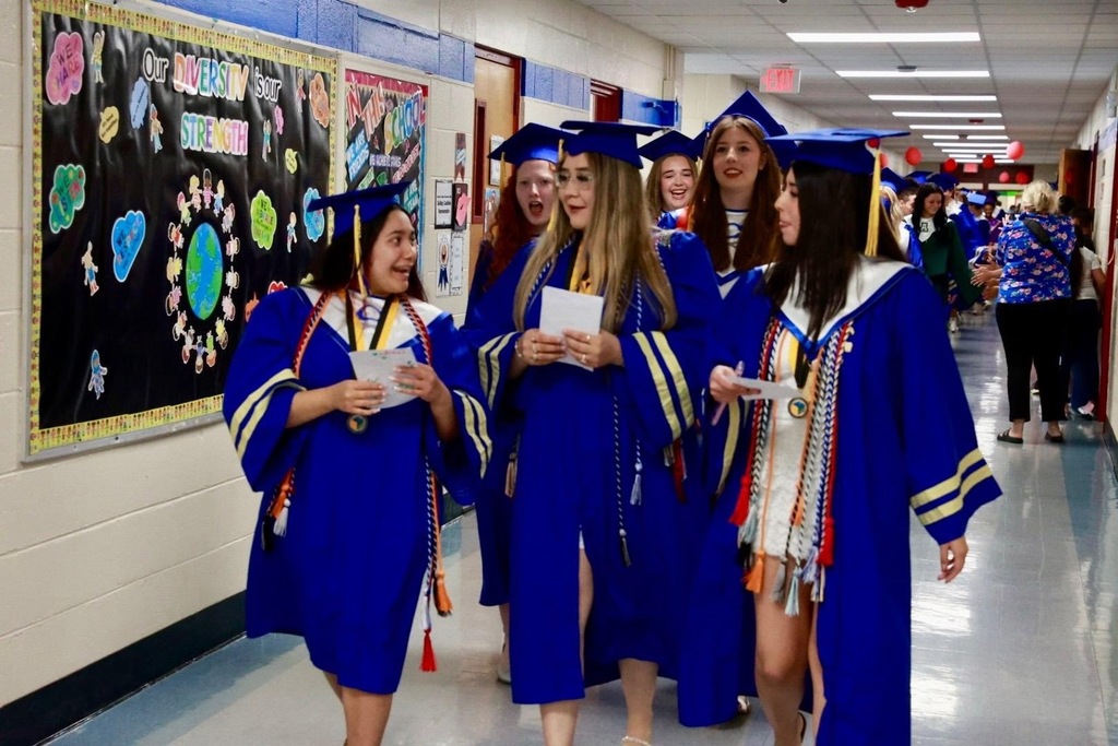 Graduates in blue gowns walk down a hallway; one holds a paper, and others hold small items.
