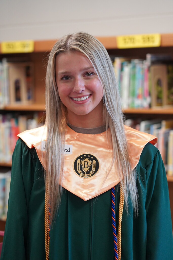 Woman with long blonde hair wearing a green and gold gown, smiling and standing in front of bookshelves.
