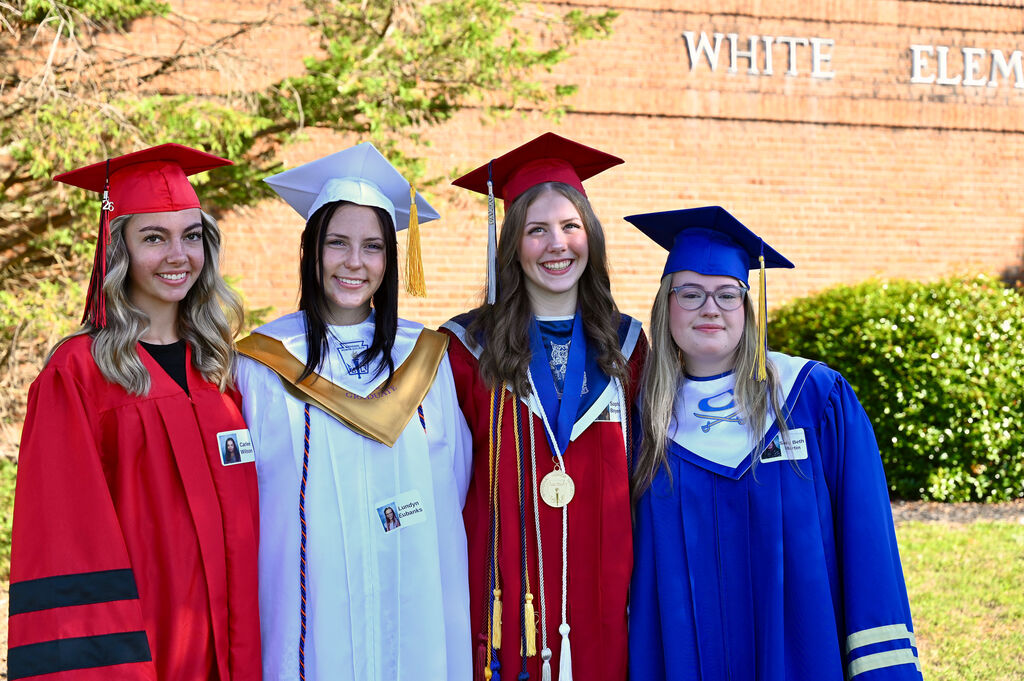 Four women in graduation gowns stand together in front of a building labeled "White Elem." They smile and look at the camera.