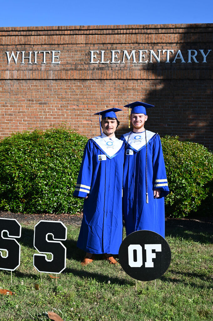 Two students in blue graduation gowns stand in front of a building with "White Elementary" written above them.