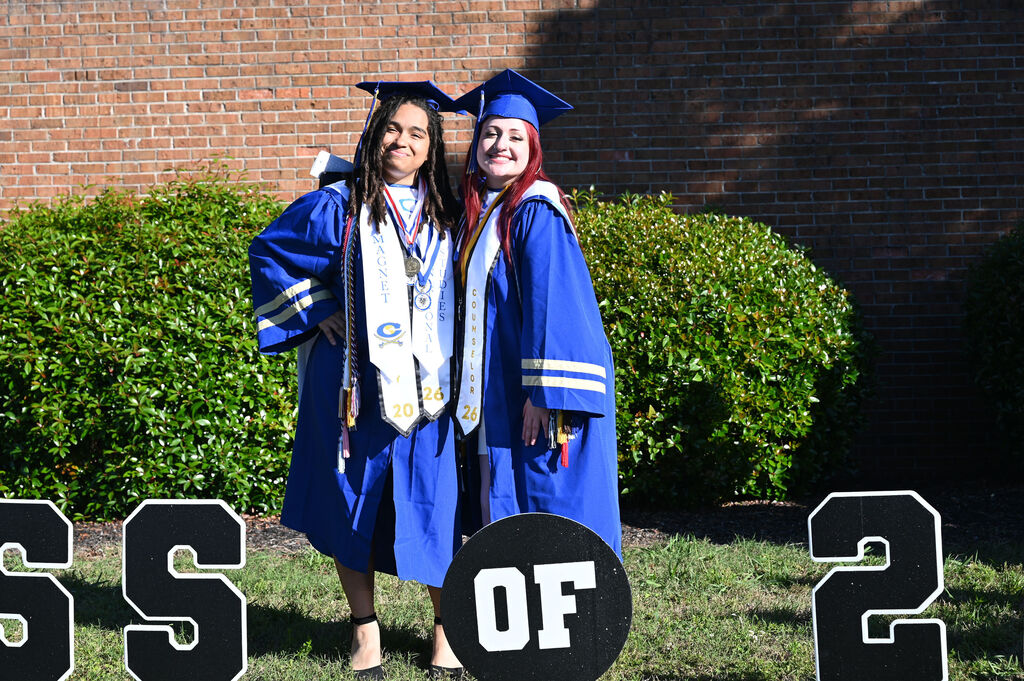 Two women in blue graduation gowns stand next to large letters on grass. One wears a cap and holds a medal.