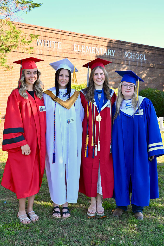 Four women in graduation gowns, posing in front of White Elementary School. Each holds a cap and gown.