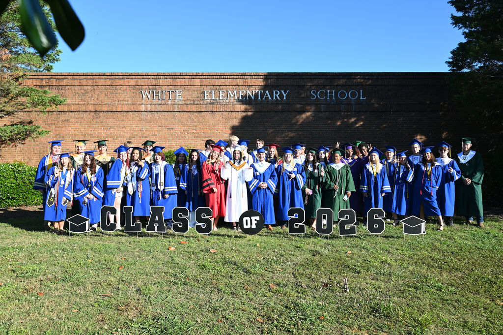 A group of people wearing caps and gowns stand in front of a school building. The sign reads "White Elementary School."