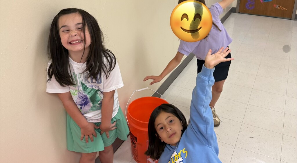 Two young girls smiling and standing in a hallway with a bucket and a smiley face.