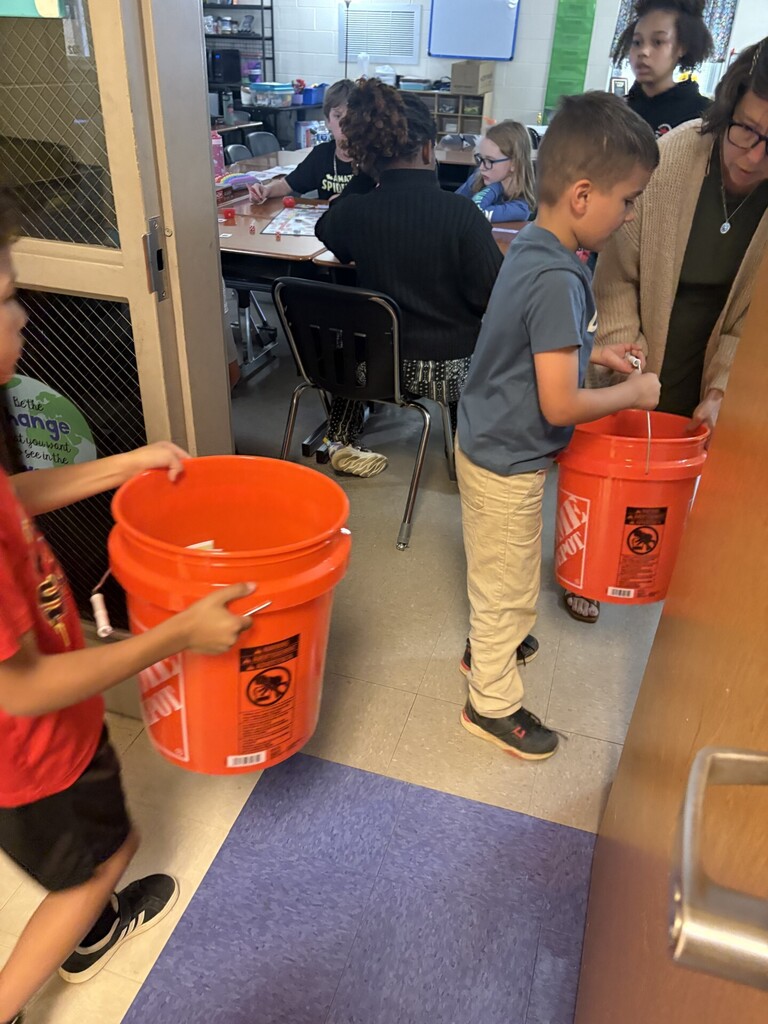 Children and adults hold orange buckets as they walk into a room. Background includes chairs and a table.