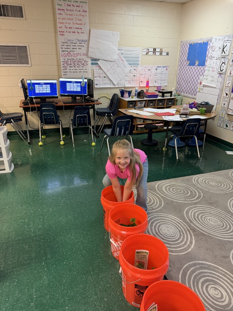 A girl crouches over a bucket filled with soil, in a classroom with green floors and white walls.