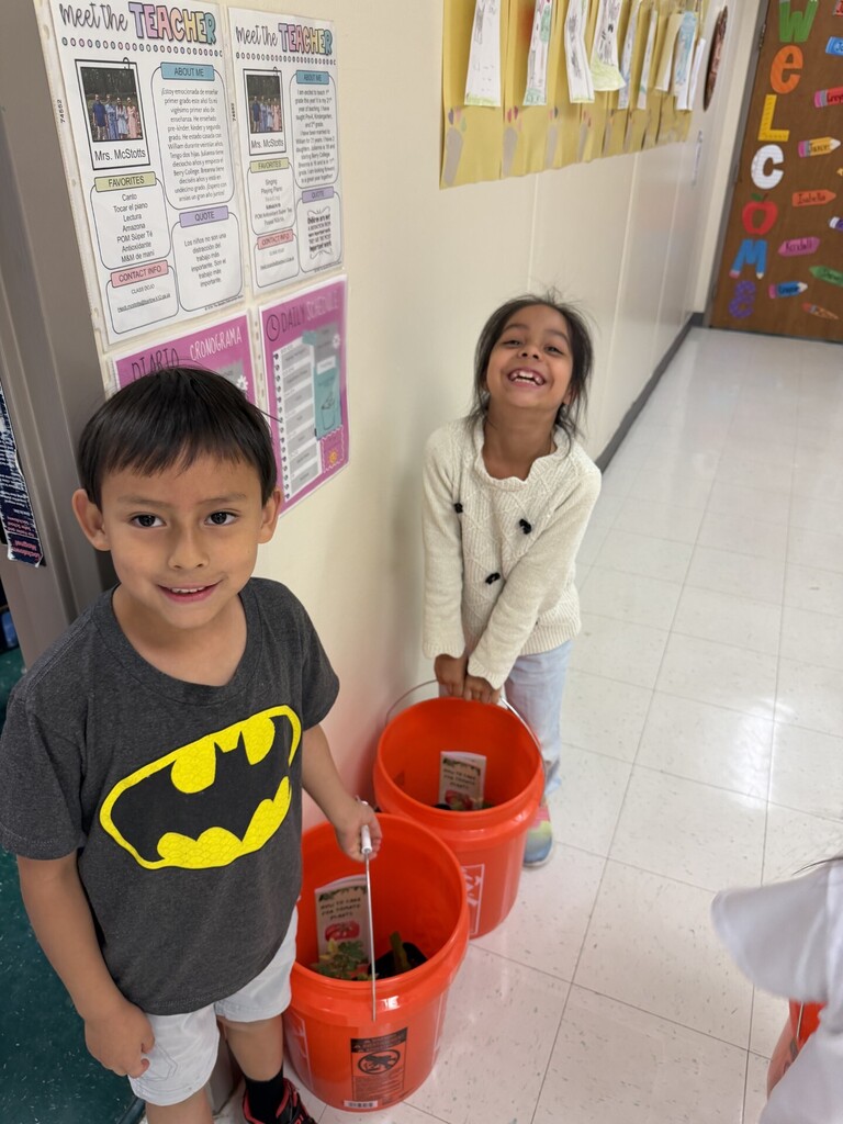 Two children in a hallway, one holding a bucket, smiling, and the other smiling with a white shirt.