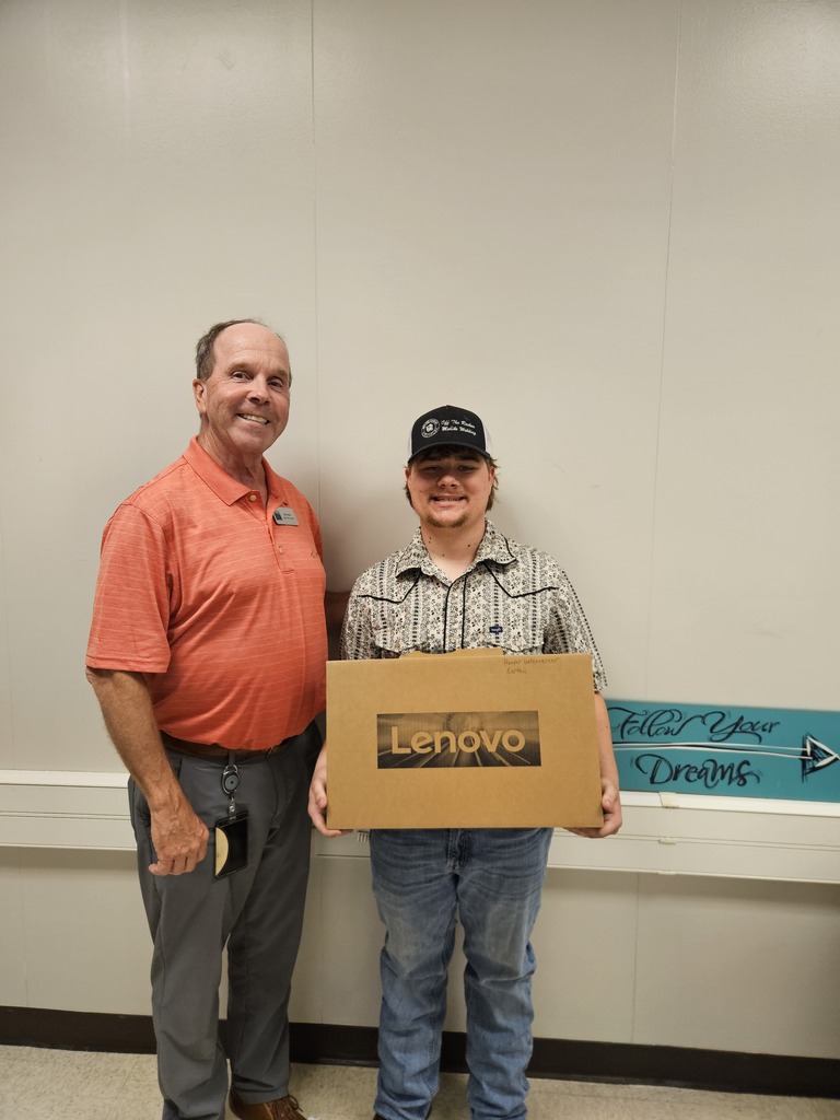 A man in an orange shirt stands next to another man in a hat holding a box with the word "Lenovo" on it. They stand in front of a white wall.