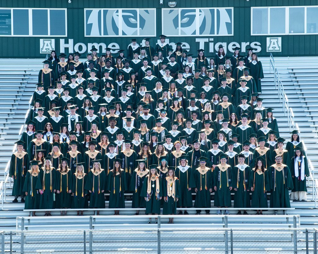 Graduating students wearing gowns and caps pose on stadium bleachers. "Home of the Tigers" is on the wall.