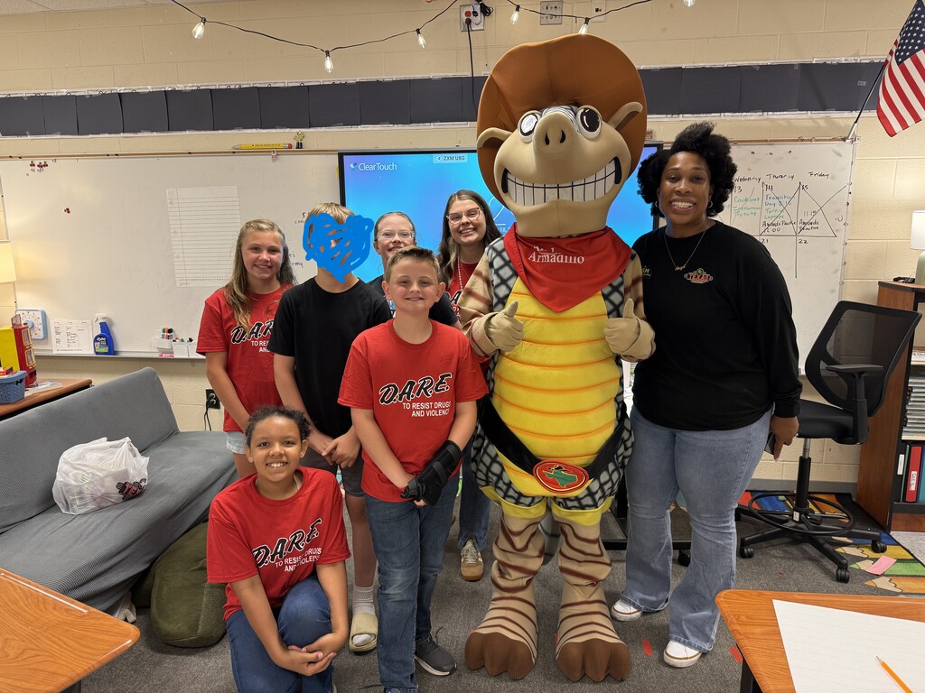 A group of children and two adults stand in a classroom, posing for a photo with a mascot.