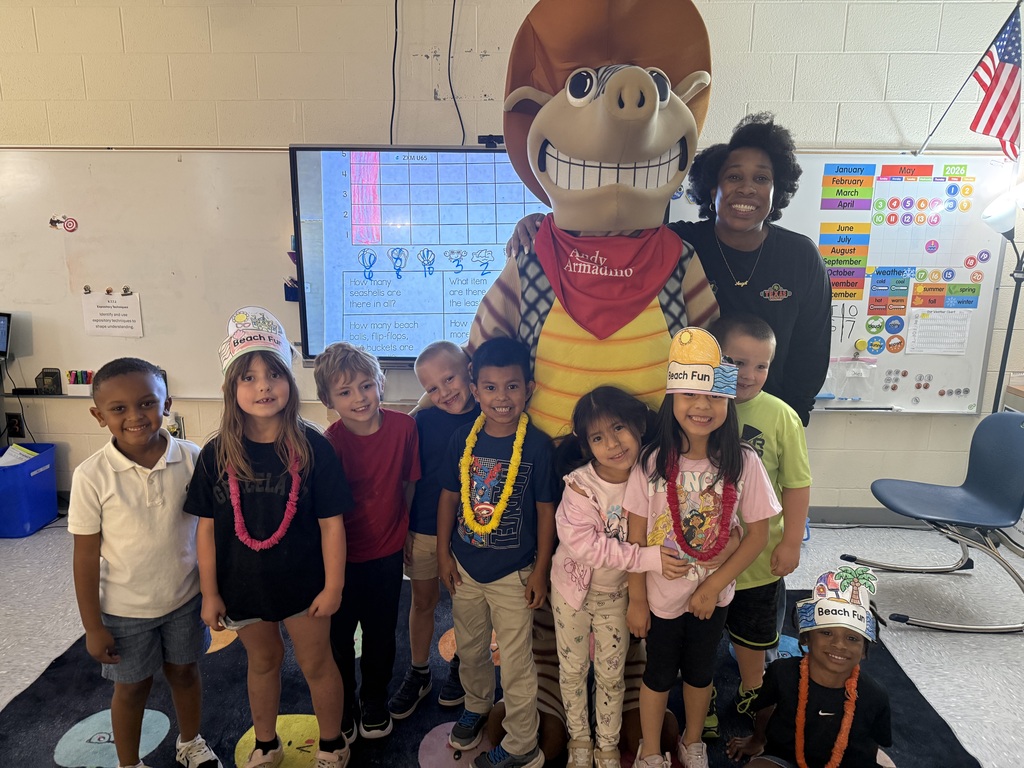 A group of children and two adults stand in a classroom. A mascot wearing a bandana is in the center.