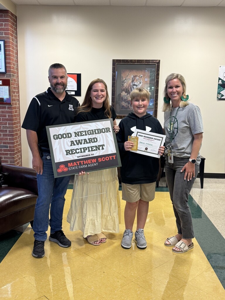 Four individuals stand in a hallway, holding a sign and awards. Two women and a boy have smiling faces.