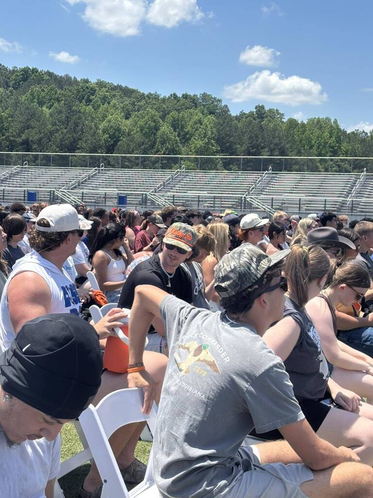 Many people sit on chairs in an outdoor stadium with bleachers, under a blue sky with some clouds.