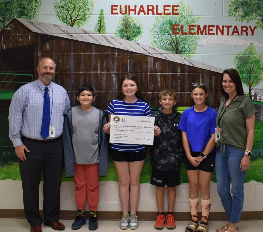 Group of children and adults posing for a photo in front of a mural with a bridge and trees.  Behind the students is a mural of a bridge with trees.  Above the mural Euharlee Elementary is written in red.  The student in the middle of the group is holding a big check made out to Euharlee Elementary for $500.  The check is signed that it is from Keep Bartow Beautiful.