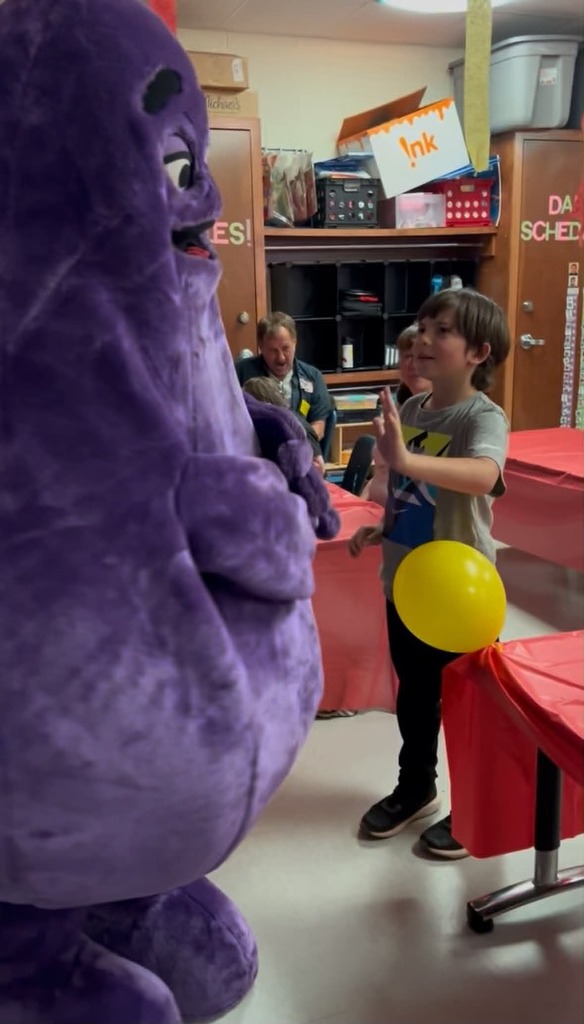 A boy waves at a large purple mascot standing next to him. Behind them, a man sits at a table.