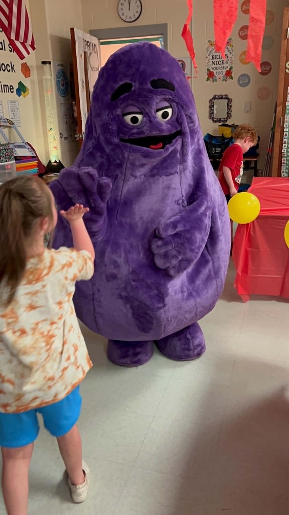 A young child interacts with a large purple mascot in a room with balloons and a table.